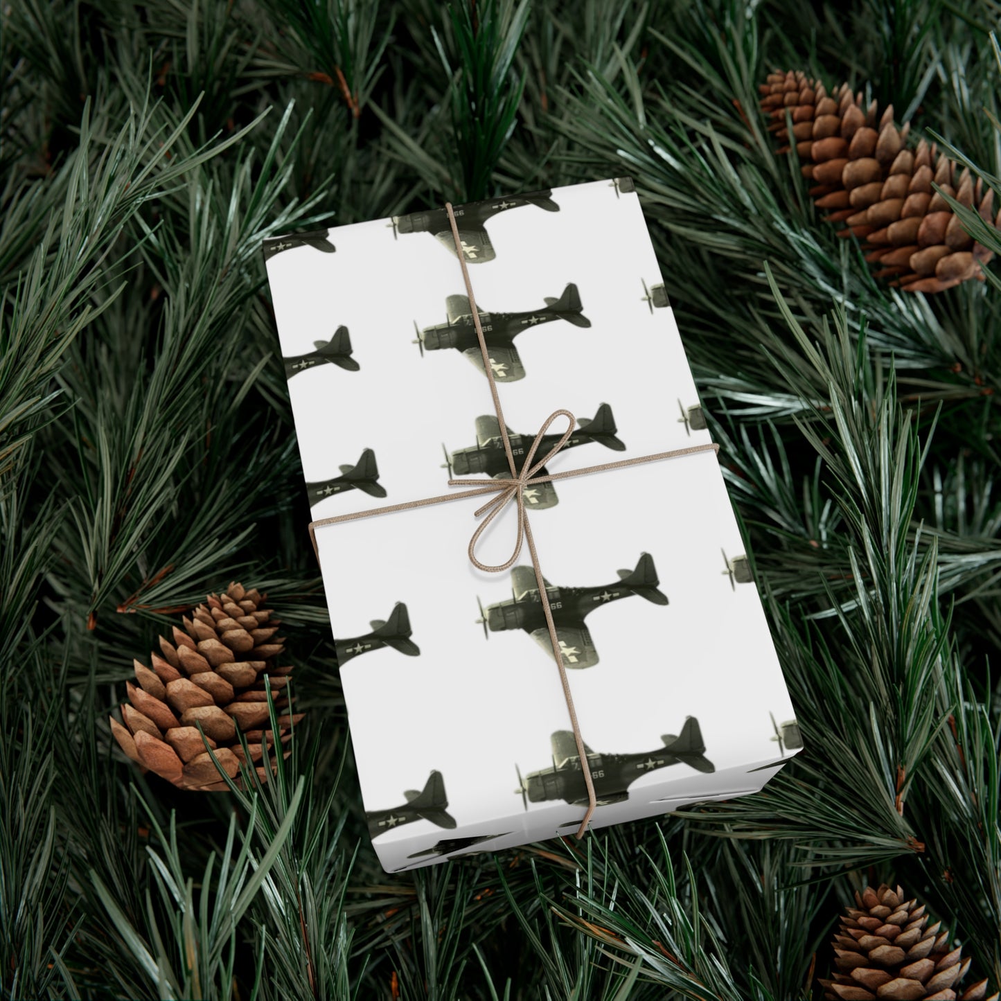 Background of pine needle branches and pine cones with a wrapped gift. Wrapping paper on the gift is white with black and white images of SBD-Dauntless dive bombers in patterned tows. The gift is tired with brown twine. 
