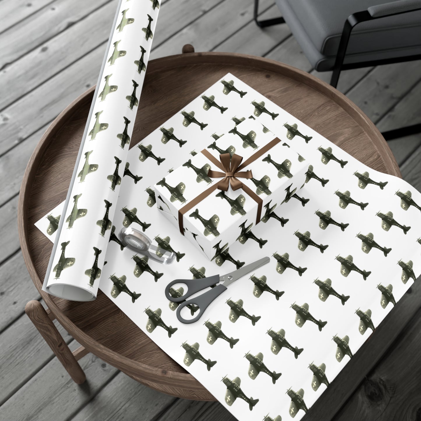 Image of hardwood floors with a round wooden table. On top of the table is a round roll of patterned SBD-Dauntless wrapping paper, a large cut out of wrapping paper, and a wrapped gift on top of it. 