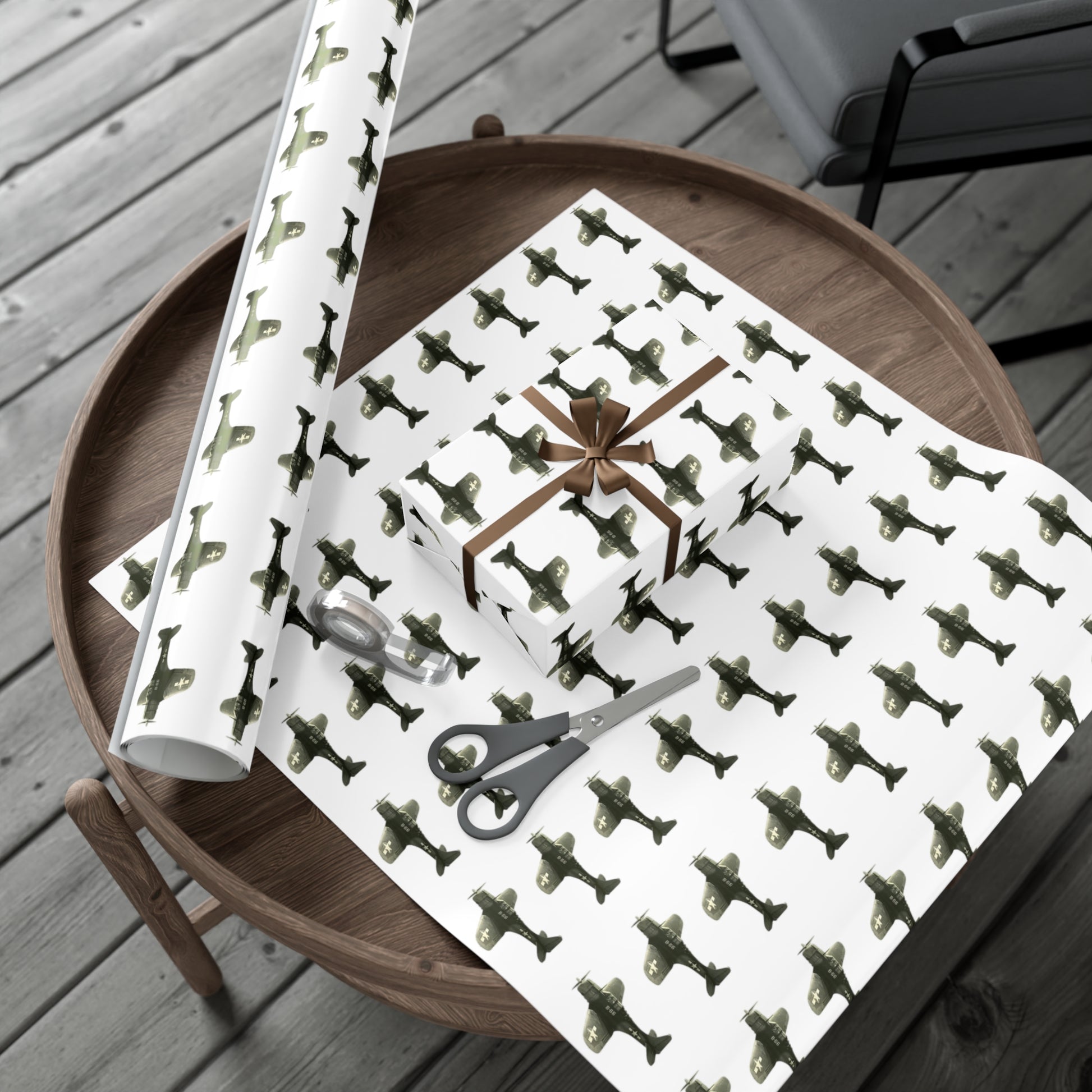Image of hardwood floors with a round wooden table. On top of the table is a round roll of patterned SBD-Dauntless wrapping paper, a large cut out of wrapping paper, and a wrapped gift on top of it. 